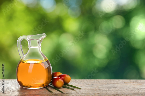 Palm oil in glass jug, tropical leaf and fruits on wooden table, space for text. Bokeh effect