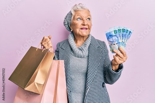 Senior grey-haired woman holding shopping bags and south africa rands banknotes smiling looking to the side and staring away thinking.