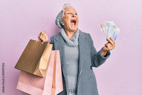Senior grey-haired woman holding shopping bags and swedish krona banknotes angry and mad screaming frustrated and furious, shouting with anger looking up.