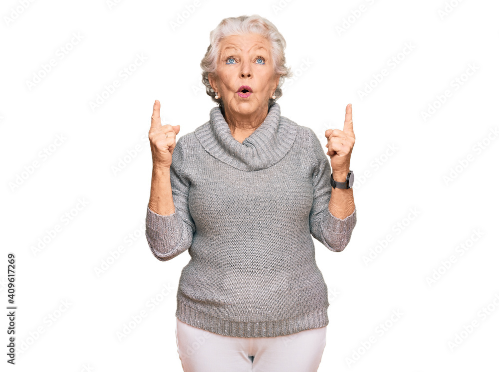 Senior grey-haired woman wearing casual winter sweater amazed and surprised looking up and pointing with fingers and raised arms. Senior grey-haired woman wearing casual winter sweater amazed and surprised looking up and pointing with fingers and raised arms.