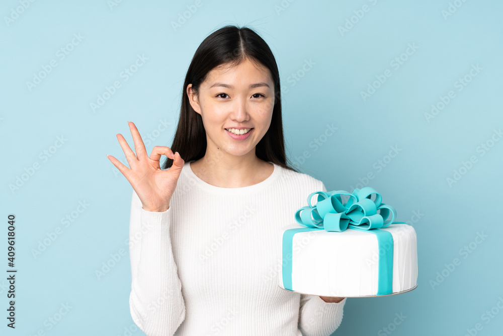 Pastry asian chef holding a big cake isolated on blue background showing ok sign with fingers Pastry asian chef holding a big cake isolated on blue background showing ok sign with fingers