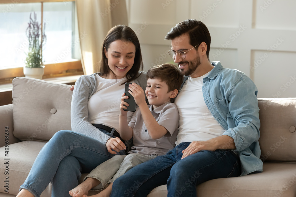 Happy young Caucasian family with small 7s son relax on sofa in living room use modern smartphone together. Smiling parents with little boy child talk on video call on cellphone. Technology concept.