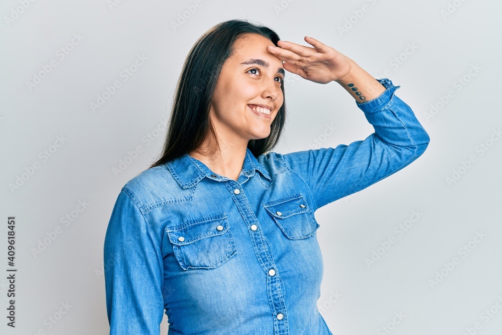 Young hispanic woman wearing casual denim jacket very happy and smiling looking far away with hand over head. searching concept. Young hispanic woman wearing casual denim jacket very happy and smiling looking far away with hand over head. searching concept.