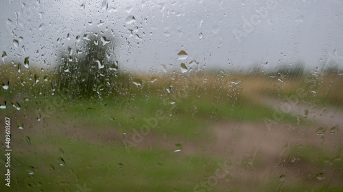 On rainy, stormy day, behind glass with sparkling raindrops, you can see blurred landscape of meadow plain with green and yellow grass, lone tree standing in distance, and muddy brown country road.