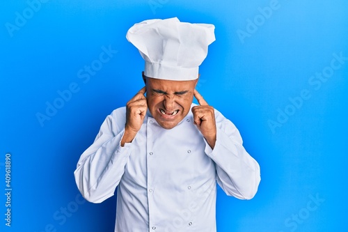 Mature middle east man wearing professional cook uniform and hat covering ears with fingers with annoyed expression for the noise of loud music. deaf concept.
