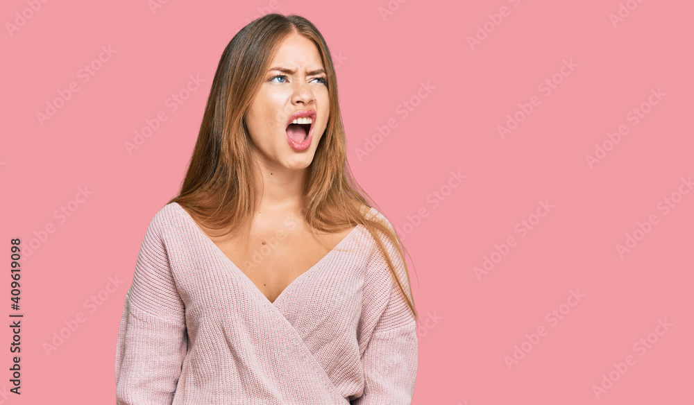 Fototapeta premium Beautiful blonde woman wearing casual winter pink sweater angry and mad screaming frustrated and furious, shouting with anger. rage and aggressive concept.