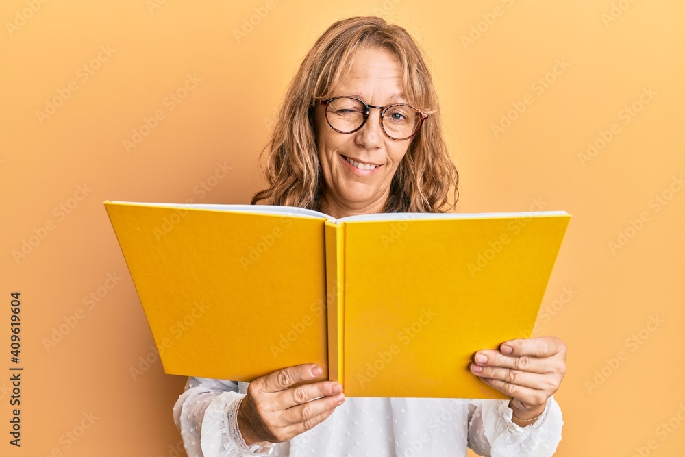 Middle age blonde woman reading a book wearing glasses winking looking at the camera with sexy expression, cheerful and happy face. Middle age blonde woman reading a book wearing glasses winking looking at the camera with sexy expression, cheerful and happy face.