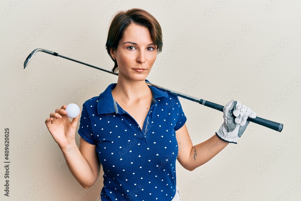 Young brunette woman with short hair holding ball and golf club relaxed with serious expression on face. simple and natural looking at the camera. Young brunette woman with short hair holding ball and golf club relaxed with serious expression on face. simple and natural looking at the camera.