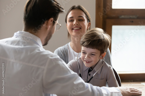 Happy small 7s boy patient have fun play with caring male pediatrician at consultation with mom in hospital. Smiling little child engaged in funny game with playful man doctor at visit to clinic.