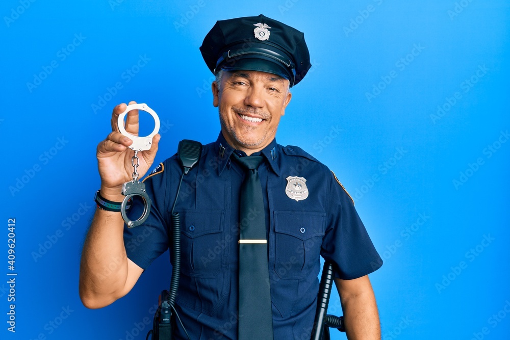 Handsome middle age mature man wearing police uniform holding metal handcuffs looking positive and happy standing and smiling with a confident smile showing teeth Handsome middle age mature man wearing police uniform holding metal handcuffs looking positive and happy standing and smiling with a confident smile showing teeth