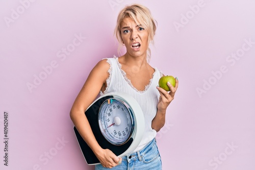 Young blonde girl holding weight machine and green apple clueless and confused expression. doubt concept.