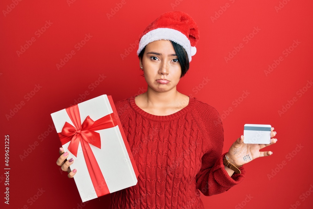Beautiful young woman with short hair wearing chrismast hat holding gift and credit card depressed and worry for distress, crying angry and afraid. sad expression. Beautiful young woman with short hair wearing chrismast hat holding gift and credit card depressed and worry for distress, crying angry and afraid. sad expression.