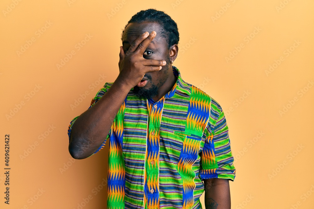 Handsome young black man wearing colorful ethnic clothes peeking in shock covering face and eyes with hand, looking through fingers afraid Handsome young black man wearing colorful ethnic clothes peeking in shock covering face and eyes with hand, looking through fingers afraid