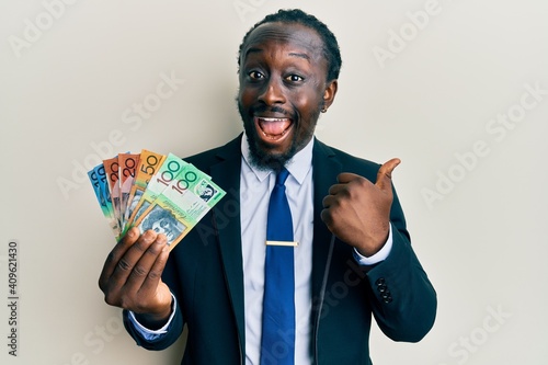 Handsome young black man wearing business suit and tie holding australian dollars pointing thumb up to the side smiling happy with open mouth