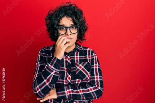Young hispanic woman with curly hair wearing casual clothes and glasses looking stressed and nervous with hands on mouth biting nails. anxiety problem. © Krakenimages.com