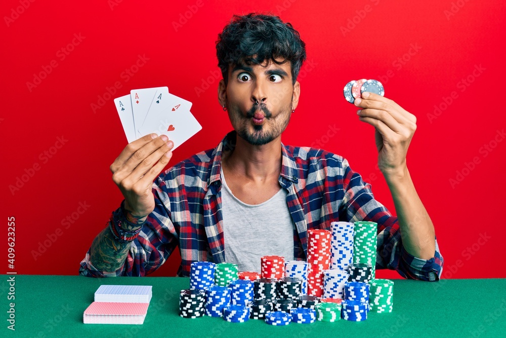 Young hispanic man playing poker holding cards and casino chips making fish face with mouth and squinting eyes, crazy and comical. Young hispanic man playing poker holding cards and casino chips making fish face with mouth and squinting eyes, crazy and comical.