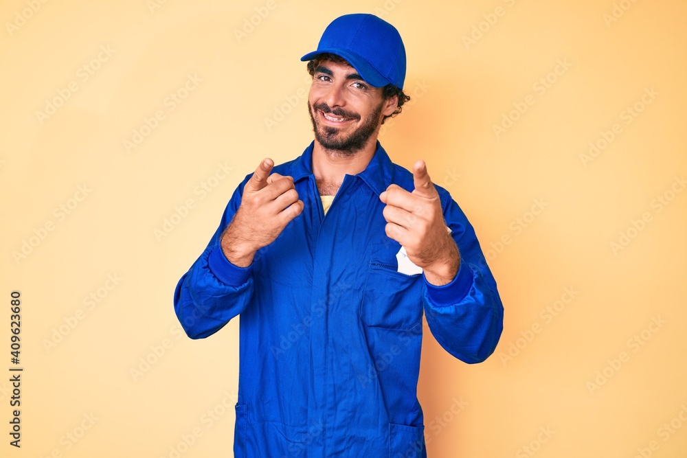 Handsome young man with curly hair and bear wearing builder jumpsuit uniform pointing fingers to camera with happy and funny face. good energy and vibes.
