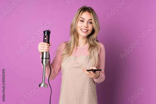 Teenager Russian girl using hand blender isolated on purple background holding coffee to take away and a mobile
