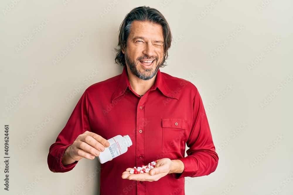 Middle age handsome man taking pills winking looking at the camera with sexy expression, cheerful and happy face. - obrazy, fototapety, plakaty 