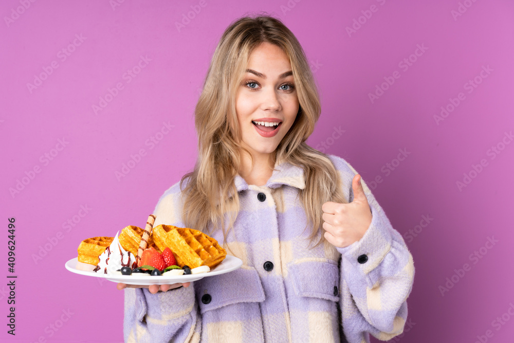 Teenager Russian girl holding waffles isolated on purple background showing ok sign and thumb up gesture Teenager Russian girl holding waffles isolated on purple background showing ok sign and thumb up gesture