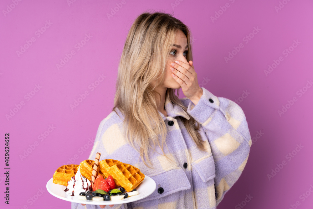 Teenager Russian girl holding waffles isolated on purple background covering mouth and looking to the side Teenager Russian girl holding waffles isolated on purple background covering mouth and looking to the side