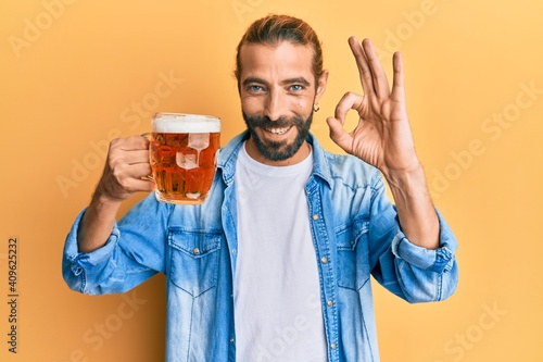 Attractive man with long hair and beard drinking a pint of beer doing ok sign with fingers, smiling friendly gesturing excellent symbol