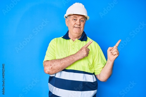 Senior grey-haired man wearing worker reflective t shirt and hardhat smiling and looking at the camera pointing with two hands and fingers to the side.