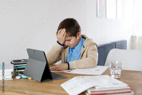 A boy is emotionally doing homework, homeschooling. He is sitting at the desk and is upset and stressed. Homeschooling in Corona. Schoolboy (7-10) years infront of a tablet. notebook. Digital learning