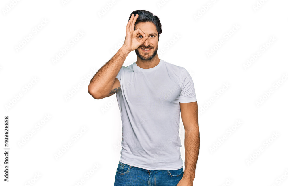 Young hispanic man wearing casual white t shirt doing ok gesture with hand smiling, eye looking through fingers with happy face. Young hispanic man wearing casual white t shirt doing ok gesture with hand smiling, eye looking through fingers with happy face.
