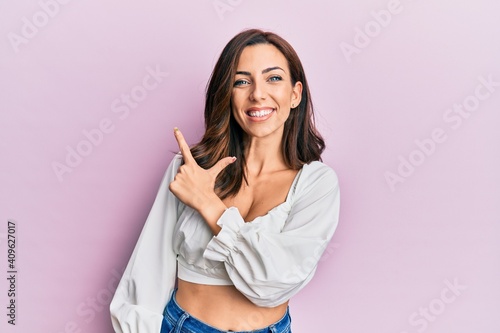 Young brunette woman wearing casual clothes over pink smiling cheerful pointing with hand and finger up to the side
