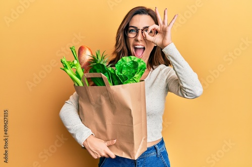 Young brunette woman holding paper bag with bread and groceries smiling happy doing ok sign with hand on eye looking through fingers
