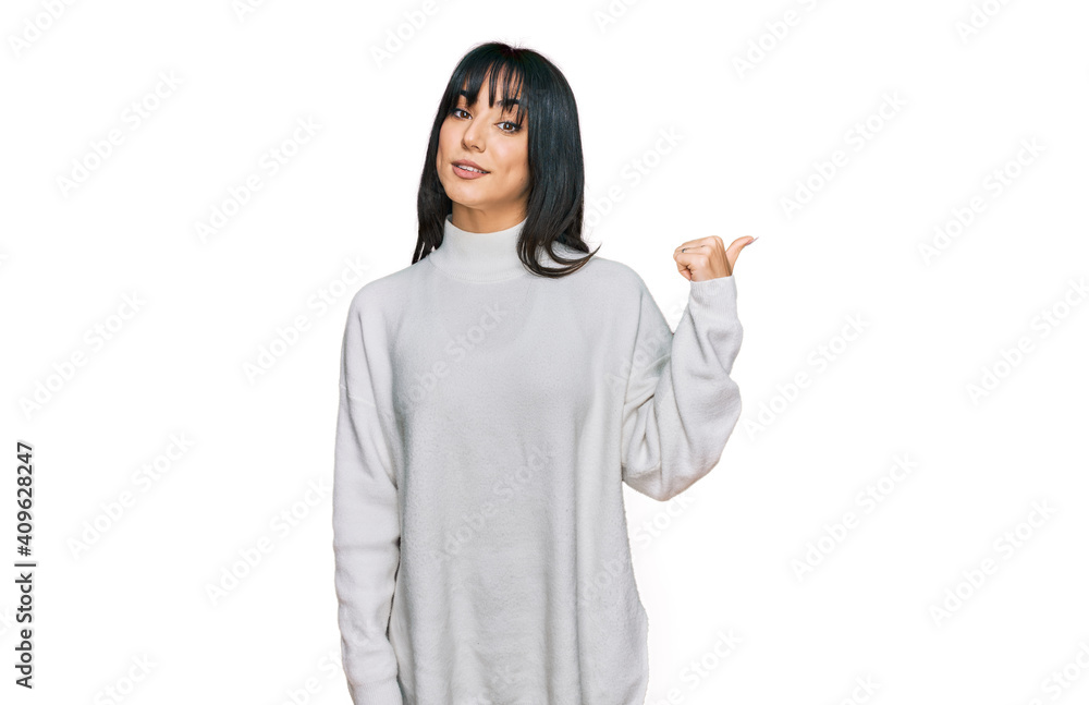 Young brunette woman with bangs wearing casual turtleneck sweater smiling with happy face looking and pointing to the side with thumb up. Young brunette woman with bangs wearing casual turtleneck sweater smiling with happy face looking and pointing to the side with thumb up.