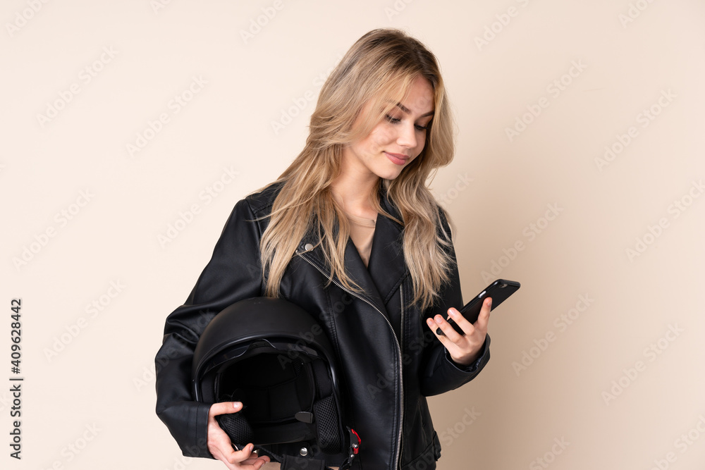 Russian girl with a motorcycle helmet isolated on beige background holding coffee to take away and a mobile Russian girl with a motorcycle helmet isolated on beige background holding coffee to take away and a mobile