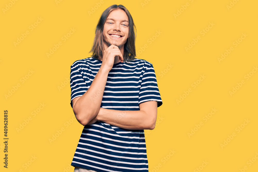 Handsome caucasian man with long hair wearing casual striped t-shirt looking confident at the camera with smile with crossed arms and hand raised on chin. thinking positive. Handsome caucasian man with long hair wearing casual striped t-shirt looking confident at the camera with smile with crossed arms and hand raised on chin. thinking positive.