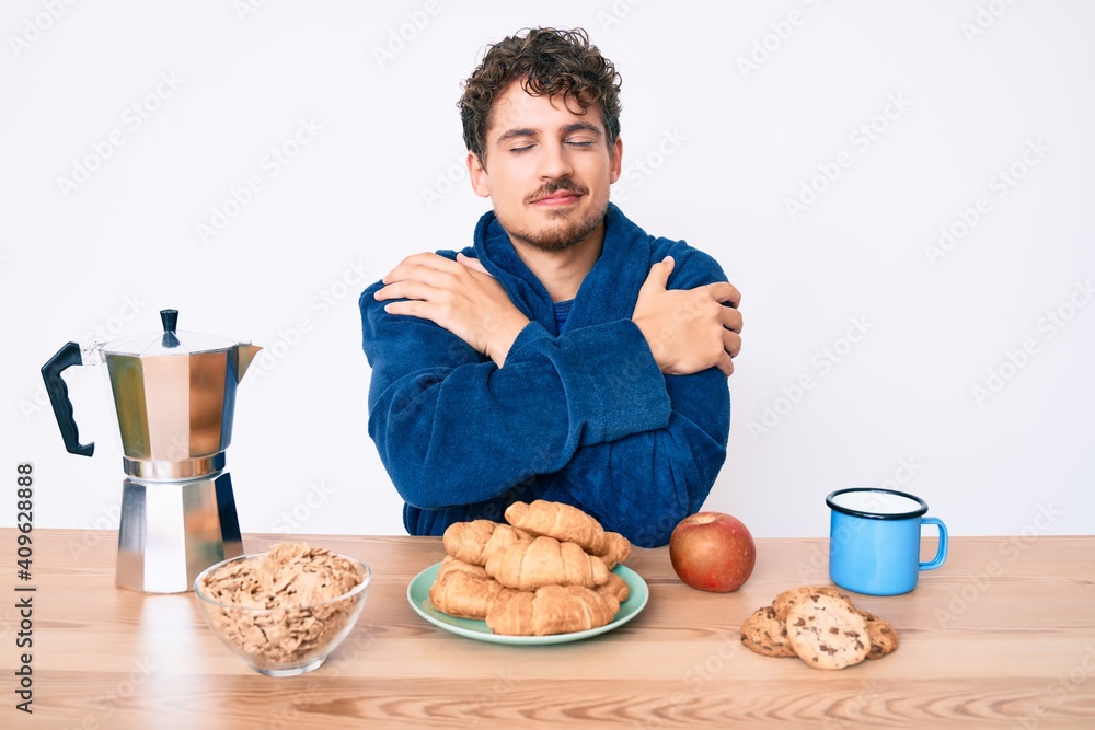 Young caucasian man with curly hair sitting on the table having breakfast hugging oneself happy and positive, smiling confident. self love and self care Young caucasian man with curly hair sitting on the table having breakfast hugging oneself happy and positive, smiling confident. self love and self care