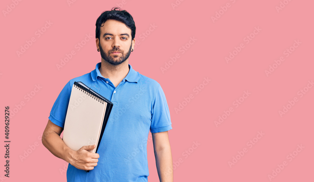 Young hispanic man holding notebook thinking attitude and sober expression looking self confident Young hispanic man holding notebook thinking attitude and sober expression looking self confident