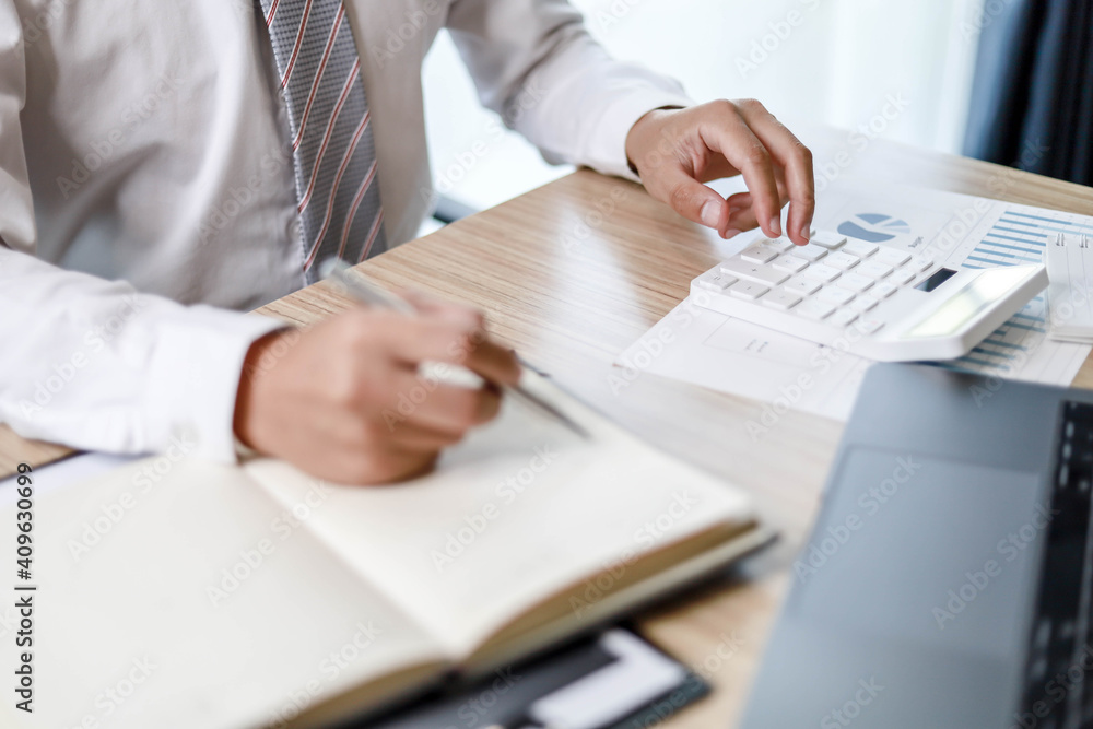 Obraz premium A male employee is taking notes in his notebook, A male employee is using a calculator and taking notes in a notebook.