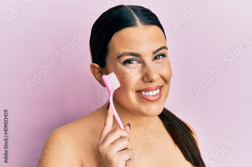 Fotografie Beautiful brunette woman using facial exfoliating brush looking positive and hap