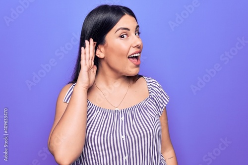 Young beautiful brunette woman wearing casual striped t-shirt standing over purple background smiling with hand over ear listening and hearing to rumor or gossip. Deafness concept.