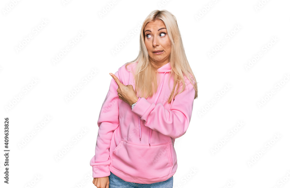 Young blonde woman wearing casual sweatshirt pointing aside worried and nervous with forefinger, concerned and surprised expression Young blonde woman wearing casual sweatshirt pointing aside worried and nervous with forefinger, concerned and surprised expression
