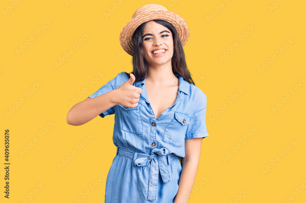 Brunette teenager girl wearing summer hat doing happy thumbs up gesture with hand. approving expression looking at the camera showing success. Brunette teenager girl wearing summer hat doing happy thumbs up gesture with hand. approving expression looking at the camera showing success.