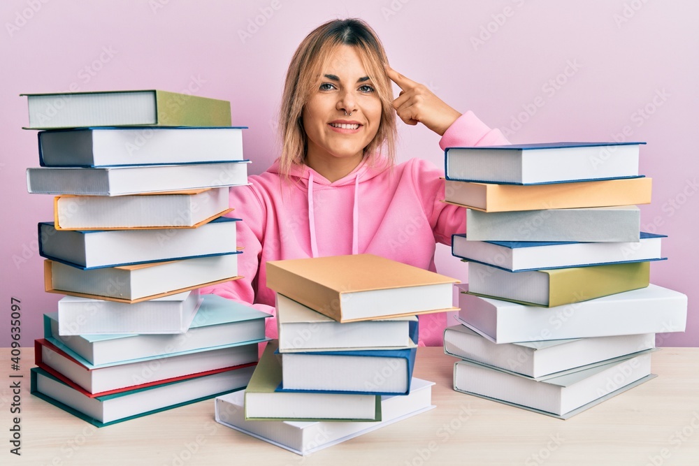 Young caucasian woman sitting on the table with books smiling pointing to head with one finger, great idea or thought, good memory Young caucasian woman sitting on the table with books smiling pointing to head with one finger, great idea or thought, good memory