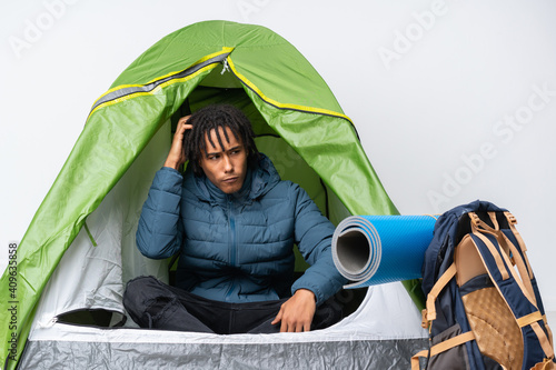 Young african american man inside a camping green tent having doubts while scratching head