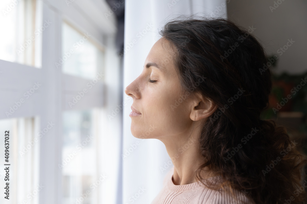 Close up head shot side view dreamy young caucasian woman breathing fresh air with closed eyes, standing near window at home. Calm peaceful millennial lady enjoying mindful morning time alone indoors.