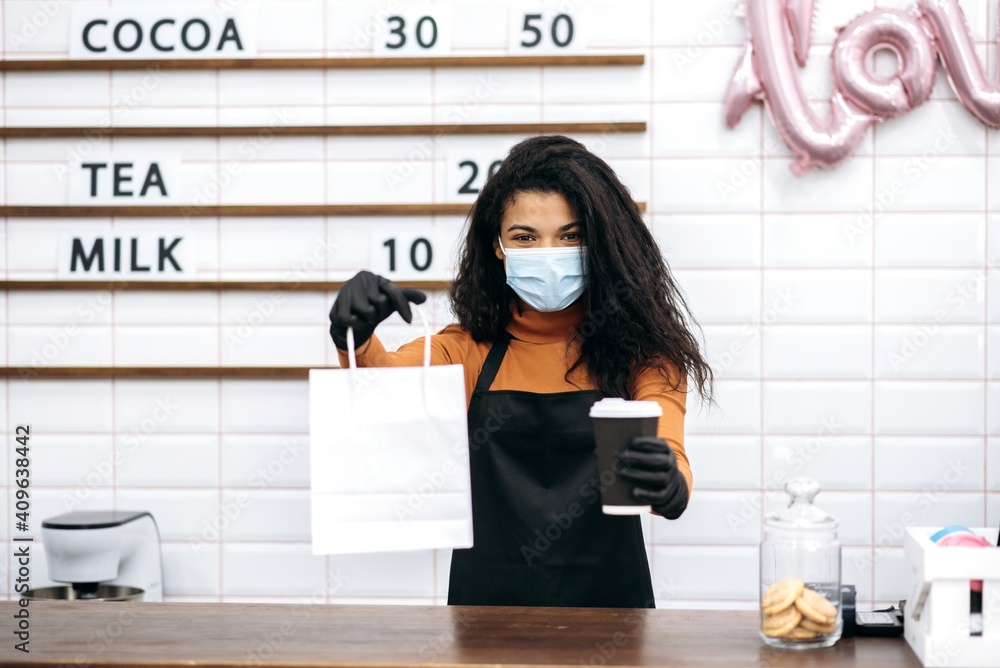 Young african american barista girl in uniform and medical mask and protective gloves gives a visitor coffee in a disposable cardboard glass and dessert in a paper bag to go. Coffee to go concept.
