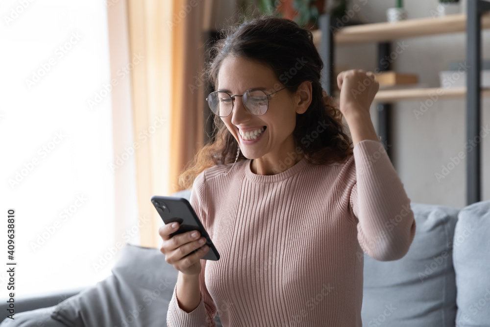 Head shot overjoyed young woman in eyewear looking at mobile phone screen, celebrating online lottery win or reading message with amazing news, feeling excited by good mobile notification at home. Head shot overjoyed young woman in eyewear looking at mobile phone screen, celebrating online lottery win or reading message with amazing news, feeling excited by good mobile notification at home.