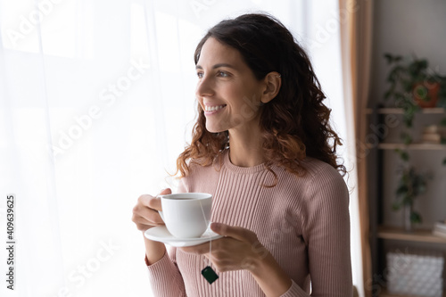 Happy beautiful young 25s caucasian dreamy woman holding cup ot hot tea, looking out of window, admiring nature or city view, enjoying peaceful morning weekend time at home or planning workday.