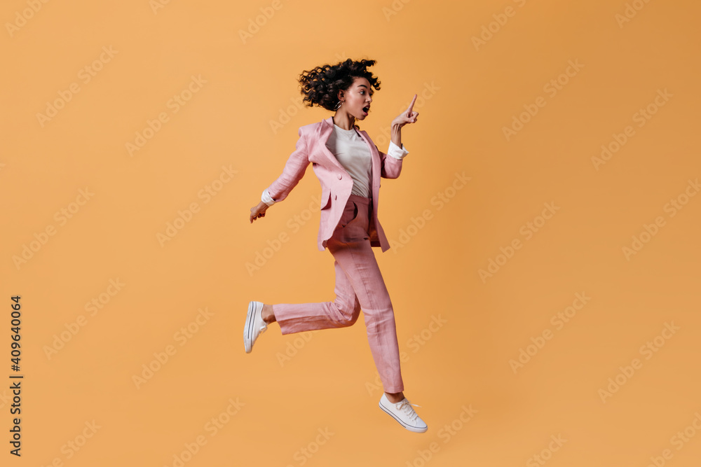 Shocked female model jumping on yellow background. Full length view of emotional mixed race woman in pink attire.