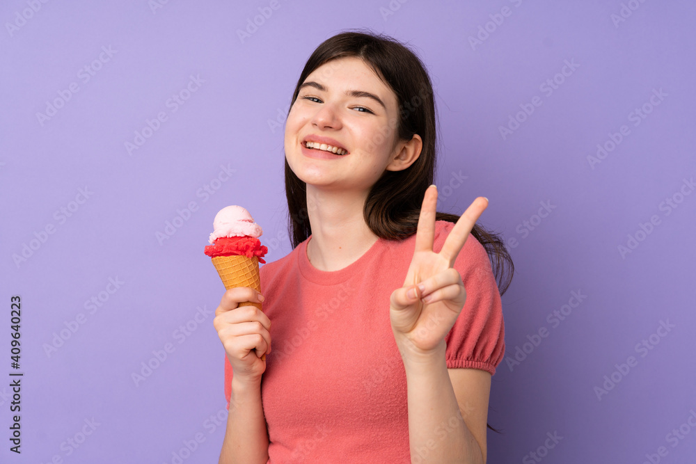 Young Ukrainian teenager girl holding a cornet ice cream over isolated purple background smiling and showing victory sign Young Ukrainian teenager girl holding a cornet ice cream over isolated purple background smiling and showing victory sign