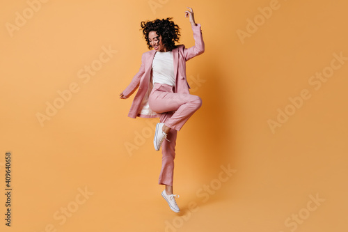 Glad curly girl jumping on yellow background. Full length view of happy lady in pink suit.